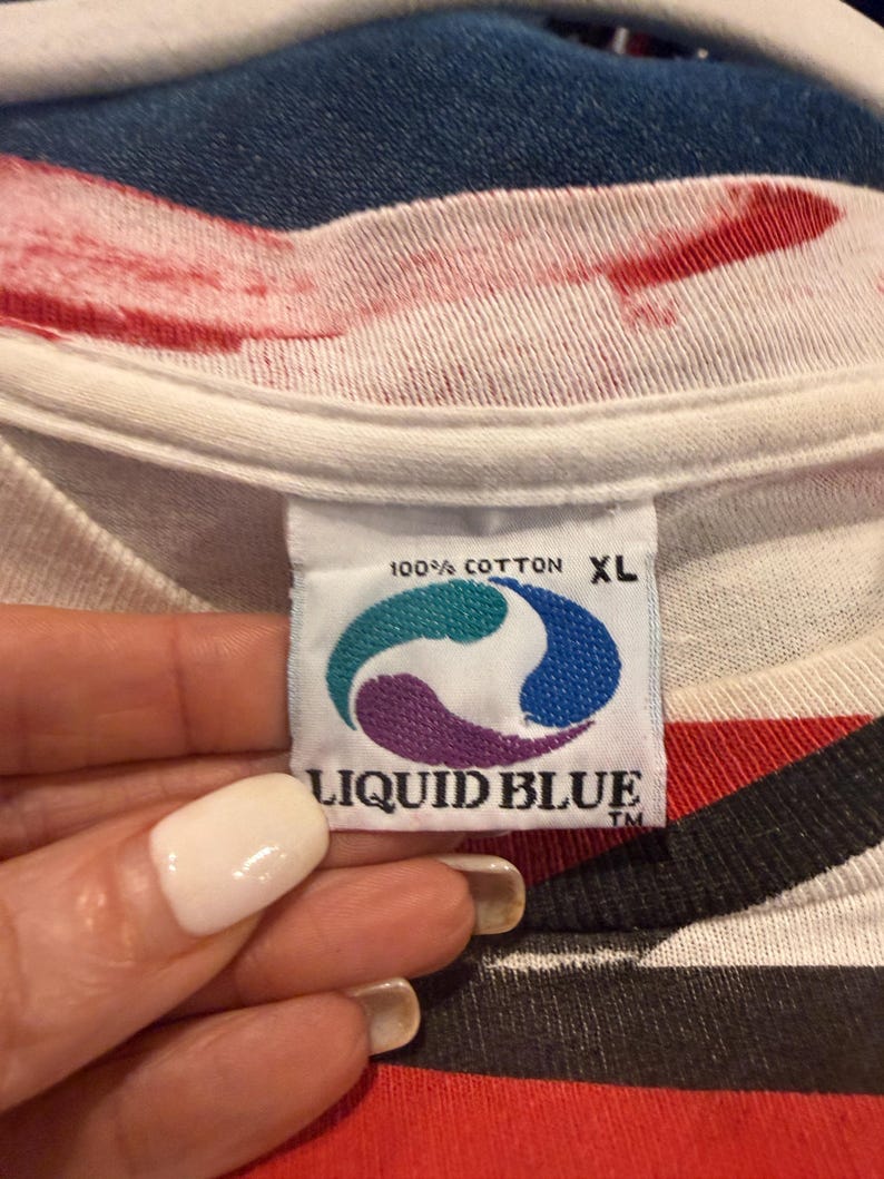 Op de afbeelding: Close-up van een wit t-shirt label met de woorden "100% COTTON XL" en het "LIQUID BLUE" logo in teal, paars en blauw. Het shirt heeft een rood en zwart gestreept ontwerp.