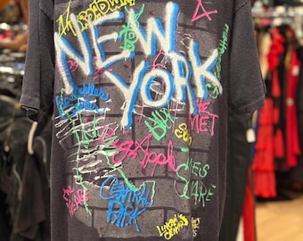 May include: Dark gray t-shirt with a colorful graffiti-style design. The design features the words "NEW YORK" in large, stylized letters, along with other text and graphics. The shirt is displayed on a mannequin.