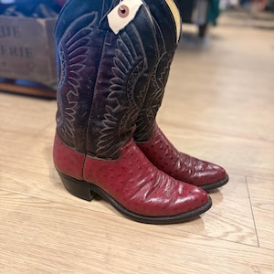 May include: A pair of cowboy boots with a two-tone design. The boots feature a deep burgundy ostrich leather foot and a black leather upper with decorative stitching. A price tag hangs from the top of the boots.