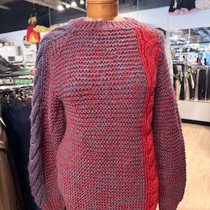 May include: A handmade sweater in shades of red, purple, and gray, displayed on a mannequin. The sweater features a cable knit design in red on the right side and a braided sleeve in purple. The body of the sweater has a textured knit.