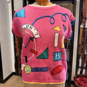 May include: A pink knit sweater vest featuring various colorful fabric appliques including geometric shapes, a blue circle with the number 2, a purple rectangle with the word "late" written in script, and a white curved banner with the text "DON'T".
