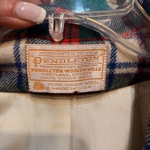May include: A Pendleton brand garment label stitched onto a plaid wool fabric with red, blue, and cream tones. The white rectangular tag features orange text reading: Warranted to be a Pendleton, Pendleton Woolen Mills, 100% Virgin Wool, Made in U.S.A.
