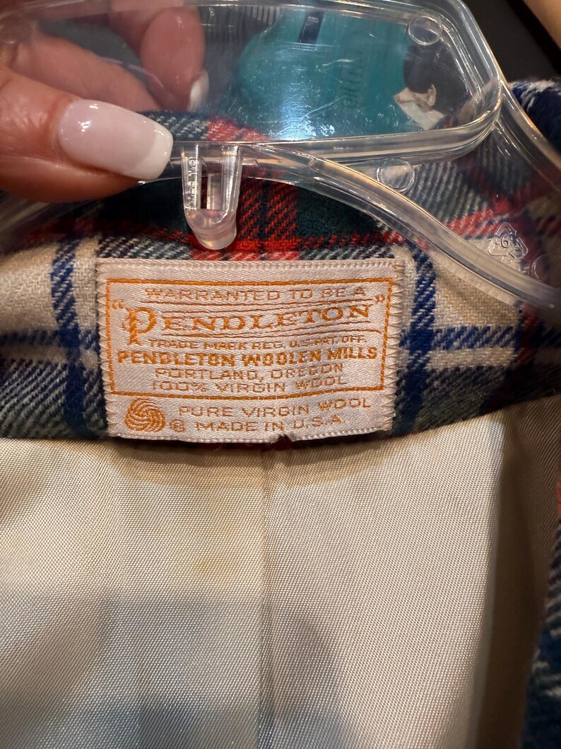 May include: A Pendleton brand garment label stitched onto a plaid wool fabric with red, blue, and cream tones. The white rectangular tag features orange text reading: Warranted to be a Pendleton, Pendleton Woolen Mills, 100% Virgin Wool, Made in U.S.A.