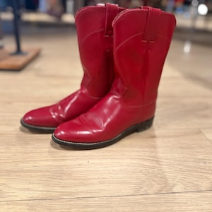 May include: A pair of bright red cowboy boots. The boots are made of a shiny material and have a classic Western design, with pointed toes and a mid-calf height. The boots are set against a light wooden floor.