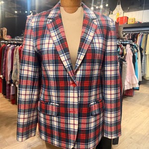 Peut inclure: Un blazer à manches longues avec un motif à carreaux rouge, bleu marine et blanc cassé. Le vêtement présente un revers cranté, deux poches à rabat sur le devant et une fermeture à deux boutons. Le tissu a un aspect texturé et est présenté sur un mannequin.