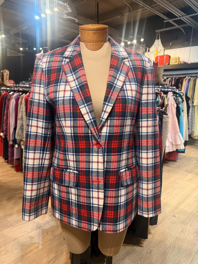 May include: A long-sleeve blazer featuring a plaid pattern in red, navy blue, and off-white. The garment has a notched lapel, two front flap pockets, and a two-button closure. The fabric has a textured appearance, and the blazer is displayed on a mannequin.