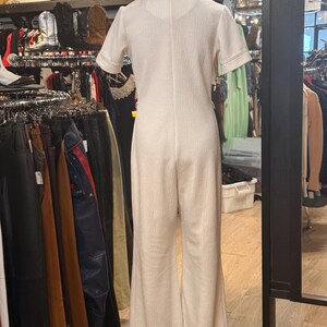 May include: Cream-colored jumpsuit with short sleeves and a collared neckline. The jumpsuit has a textured fabric and flared legs. The sleeves and collar are trimmed with a contrasting stitch. The garment is displayed on a mannequin in a vintage clothing store.