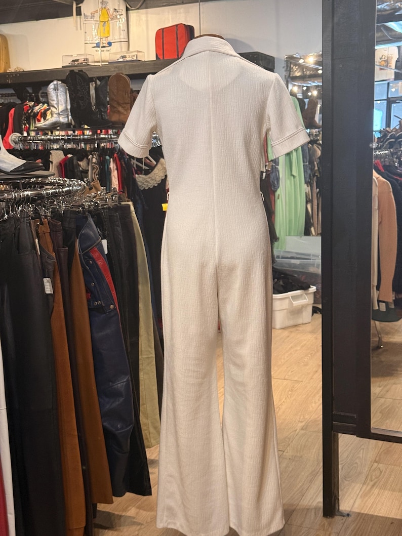 May include: Cream-colored jumpsuit with short sleeves and a collared neckline. The jumpsuit has a textured fabric and flared legs. The sleeves and collar are trimmed with a contrasting stitch. The garment is displayed on a mannequin in a vintage clothing store.