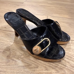 Può includere: Un paio di sabot in vernice nera con suola a piattaforma e tacco in legno. Ogni scarpa presenta un'ampia fascia sulla parte superiore decorata con una fibbia dorata e un emblema circolare beige con il logo CC intrecciato al centro.