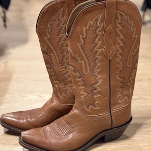 May include: A pair of brown leather cowboy boots. The boots have pointed toes, decorative stitching, and a classic Western design. The boots are made of a light brown leather and have a worn appearance. The boots are on a wooden surface.