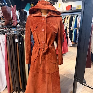 Pode incluir: Um casaco de camurça cor de ferrugem com capuz, fechos de botão e cinto de amarrar. O casaco tem dois bolsos frontais e uma saia longa e rodada. O casaco é exibido em um manequim em uma loja de roupas vintage.