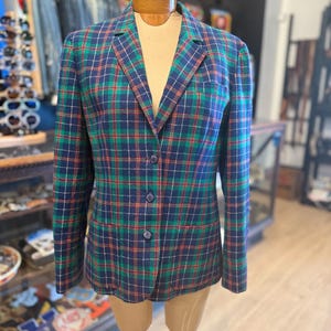 May include: A vintage plaid blazer in navy blue, green, and red, displayed on a mannequin. The blazer features a classic plaid pattern, a notched collar, and three button closure. The jacket is a classic piece of vintage clothing.