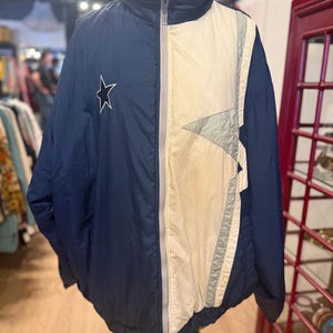 May include: A vintage navy blue and cream-colored jacket with a full zipper closure. The jacket features a white star on the left chest and a color-blocked design with gray accents. The jacket is displayed on a mannequin.