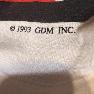 Op de afbeelding: Close-up van een witte stof met rode en zwarte strepen. De tekst "&copy; 1993 GDM INC." is in het zwart gedrukt. De stof lijkt een gebreid materiaal te zijn.