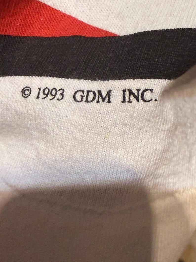 Op de afbeelding: Close-up van een witte stof met rode en zwarte strepen. De tekst "&copy; 1993 GDM INC." is in het zwart gedrukt. De stof lijkt een gebreid materiaal te zijn.