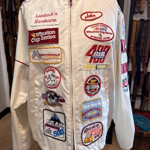 May include: A white zip-up racing jacket featuring numerous embroidered patches including Leinbach's Hardware, Winston Cup Series, Martinsville Speedway, Rebel 500, Peak Performance, John, Dover Downs, RIR 400, and Darlington Raceway logos.