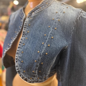 May include: A denim bolero jacket in a medium wash, adorned with small, gold-colored studs. The jacket features a collarless design and three-quarter sleeves. The garment is displayed on a mannequin.
