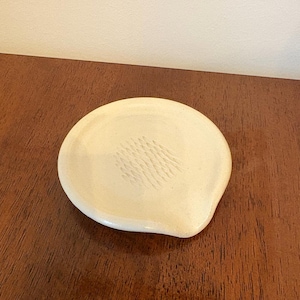 May include: A small, off-white ceramic spoon rest with a textured pattern in the center. The spoon rest has a rounded shape with a slight indentation on one side. The background is a dark wood surface.