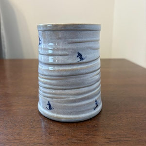May include: A cylindrical ceramic mug with a light beige, glossy glaze and a series of horizontal ridges along the body. The mug is decorated with small, dark blue stamped silhouettes of skiers in various poses scattered across the exterior surface.