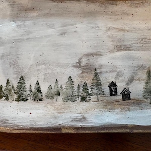 May include: A rectangular, rustic wooden tray with a winter scene painted on the surface. The design features a snowy landscape with evergreen trees and two small houses with smoke rising from the chimneys. The tray has a white and gray background.