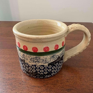 May include: A ceramic mug with a cream-colored base featuring a row of red dots near the rim, a dark green horizontal band, and a lower section decorated with a dark blue pattern of small circles. The mug has a textured, braided-style handle.