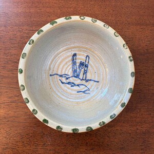 May include: A round ceramic bowl with a light beige speckled glaze. The rim features a pattern of small green dots. The interior base displays a blue hand-painted graphic of two vertical shapes resembling skis or poles above a wavy line.