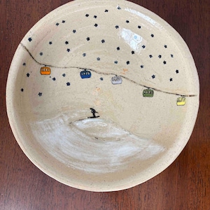 May include: A beige ceramic bowl with a hand-painted winter scene. The bowl features a skier on a snowy slope, a ski lift with colorful gondolas, and dark blue star-like accents. The bowl is round and has a slightly raised edge.