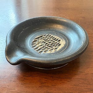 May include: A black ceramic grater with a textured grating surface in the center. The grater has a small spout on one side and a white base. The kitchen tool is designed for grating ingredients.
