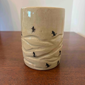 May include: A cylindrical ceramic mug with a speckled beige finish and a textured surface featuring horizontal, wave-like ridges. The mug is decorated with small, black, hand-painted silhouettes of skiers in various poses across the exterior.