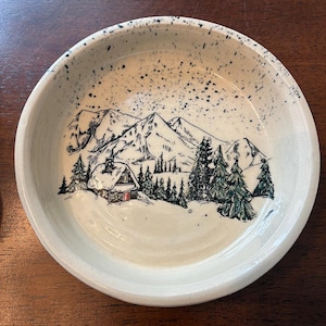 Puede incluir: Un cuenco redondo de cerámica blanquecina con una escena invernal dibujada a mano. La ilustración presenta una cabaña con puerta roja, árboles de hoja perenne y montañas nevadas. El borde tiene un patrón moteado.