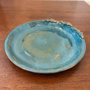 May include: A round, teal-colored ceramic dish with a glossy finish. The surface features a subtle, grid-like texture imprinted in the center. The rim is uneven and decorated with a metallic gold-toned accent along one side of the edge.
