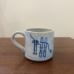 May include: A light gray ceramic mug with a speckled texture and a single handle. The front of the mug features a dark blue hand-painted line drawing depicting a pair of skis and ski poles arranged in a stylized, abstract composition.