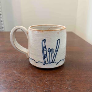 May include: A cream-colored ceramic mug with a speckled texture and a brown rim. The mug features a hand-drawn blue illustration of two skis standing upright in the snow. It has a single curved handle on the left side and a cylindrical body.