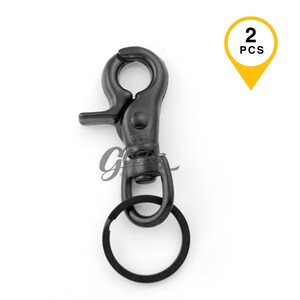 May include: A matte black metal swivel lobster claw clasp with a spring-loaded trigger mechanism and an attached circular split key ring. A yellow location pin icon in the top right corner contains the black text 2 PCS, indicating a set of two.