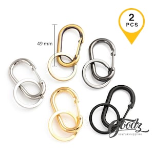 May include: Five oval carabiner clips with attached round split rings in silver, gold, gunmetal, and black finishes. One gold clip is labelled with a height of 49 mm. A yellow pin icon in the top right corner displays the text 2 PCS, and the bottom right corner features the text Goodz craft & supplies.