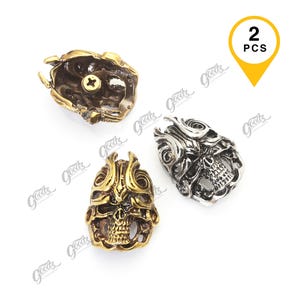 May include: A set of two metal skull-shaped beads or charms, one in a gold tone and one in a silver tone. Each piece features intricate, raised scrollwork patterns and a hollowed-out design. The gold-toned piece shows a screw head on its underside.