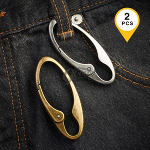 May include: Two oval-shaped metal carabiner clips, one in a silver tone and one in an antique brass tone, featuring a spring-loaded gate mechanism. The silver carabiner is attached to a belt loop on dark denim fabric. A yellow tag displays 2 PCS.