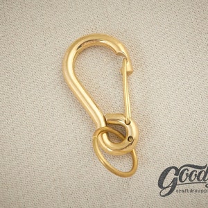 Puede incluir: Un mosquetón de metal dorado con un cierre de gatillo y una abertura redondeada. El mosquetón está diseñado para sujetarse a llaveros, bolsos u otros artículos.