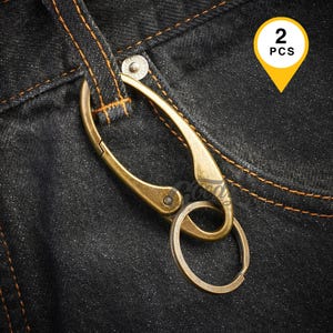 May include: A set of two antique brass-toned metal carabiner keychains with a spring-loaded gate and an attached circular split ring. The carabiner is hooked onto a dark denim fabric loop with orange stitching and a silver-colored metal rivet.