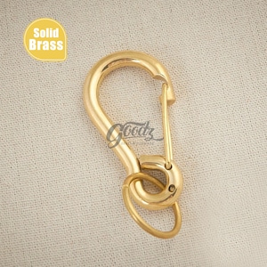 May include: A solid brass carabiner clip with a spring-loaded gate and a small attached ring. The clip is gold-colored and features the text "Solid Brass" in a yellow oval. The background is a neutral fabric.