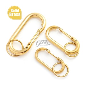May include: Three solid brass carabiner clips in varying sizes. Each clip has a spring-loaded gate and a small ring. The upper left corner of the image has a yellow oval with the words "Solid Brass". The clips are on a white background.