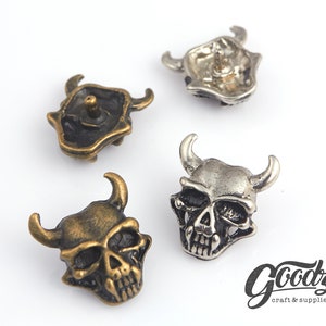 Puede incluir: Cuatro tachuelas metálicas con forma de calavera con cuernos. Las tachuelas tienen varios acabados, como latón antiguo y plata. Las calaveras tienen detalles y están diseñadas para proyectos de artesanía y suministros. El logotipo de Goodz craft & supplies está en la esquina inferior derecha.