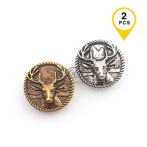 May include: Two metal buttons featuring deer head designs. One button is bronze-coloured with a mountain and forest background, and the other is silver-coloured. The buttons are round with a rope-like border. The image includes a yellow tag that says "2 PCS".