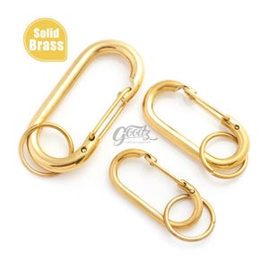 May include: Three oval-shaped solid brass carabiner clips of varying sizes, each featuring a spring-loaded gate and an attached circular split ring. The metal has a polished gold-tone finish. Text on the image reads Solid Brass and Goodz.
