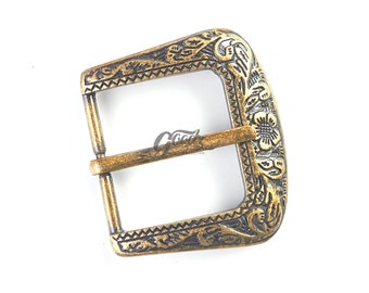 May include: A brass-toned metal belt buckle featuring an intricate embossed floral and scroll pattern along the frame. The buckle has a rectangular shape with rounded corners and a central prong, displaying a weathered finish with dark recessed details.
