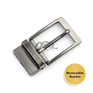 May include: A rectangular gunmetal belt buckle with a brushed metallic finish and a central prong. The buckle features a swivel mechanism for reversible use. A yellow circular graphic in the corner displays the text Reversible Buckle.