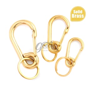 May include: Three solid brass carabiner clips in varying sizes. The clips have a shiny gold finish and are shown with key rings. The image includes a yellow label that reads "Solid Brass".