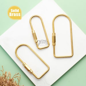 May include: Three solid brass keychains in a U-shape design. The keychains have a rectangular centre piece with a screw closure. The keychains are displayed on a white surface with a green background. The text "Solid Brass" is in a yellow box.