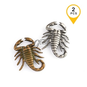 May include: Two scorpion-shaped metal charms, one in a bronze finish and one in a silver finish. Each scorpion features detailed segmented bodies, pincers, and curved tails. A yellow graphic in the top right corner displays the text 2 PCS.