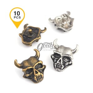 Puede incluir: Cuatro tachuelas decorativas de metal con forma de calavera y cuernos. Las tachuelas son en tonos oro antiguo y plata. La imagen incluye una etiqueta amarilla que dice "10 PCS" y el logotipo de Goodz craft & supplies.